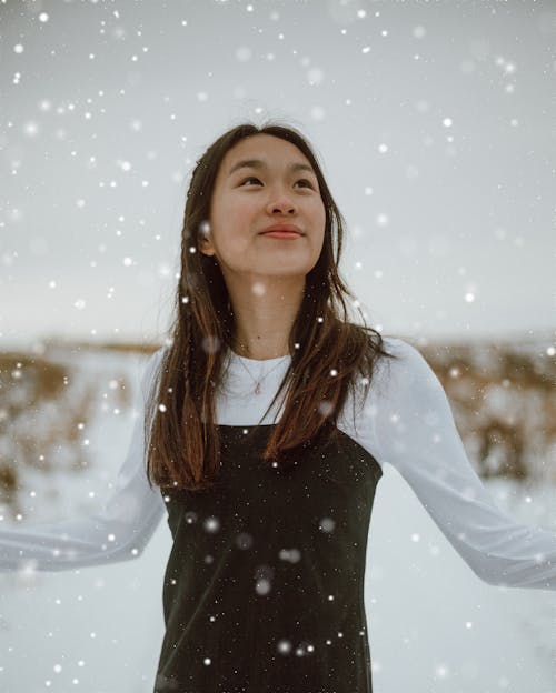 Young cheerful ethnic female in casual outfit admiring snowfall while looking up in wintertime