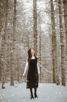 Young ethnic female in casual apparel and boots looking up on walkway among dry trees in snowfall