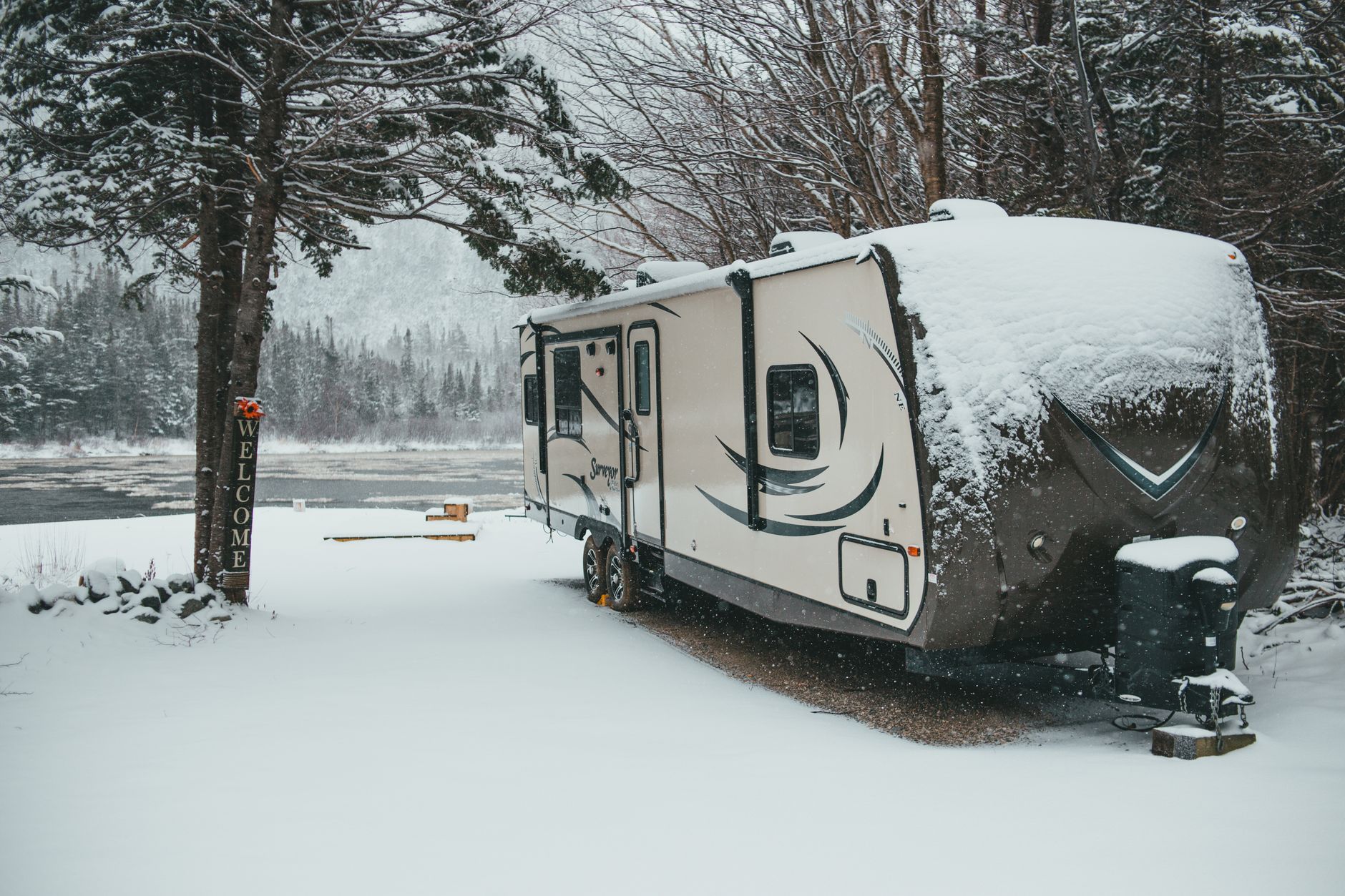 Travel trailer parked and covered in fresh snow beside a frozen lake, stored for winter