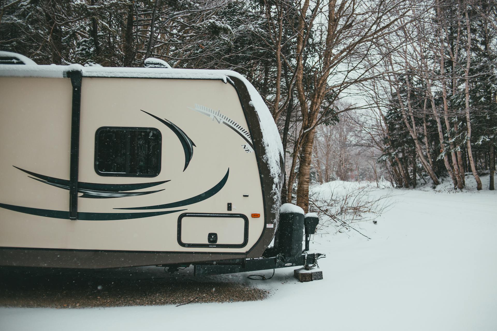 Camper trailer parked in coniferous and deciduous forest on road trip in snowfall in gloomy weather in winter in daytime