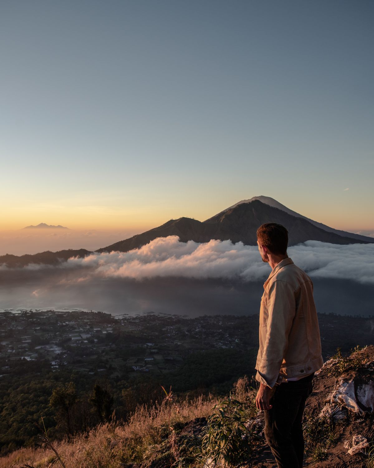 A hiker standing on a summit ridge at sunrise, above a sea of cloud with a volcano on the horizon