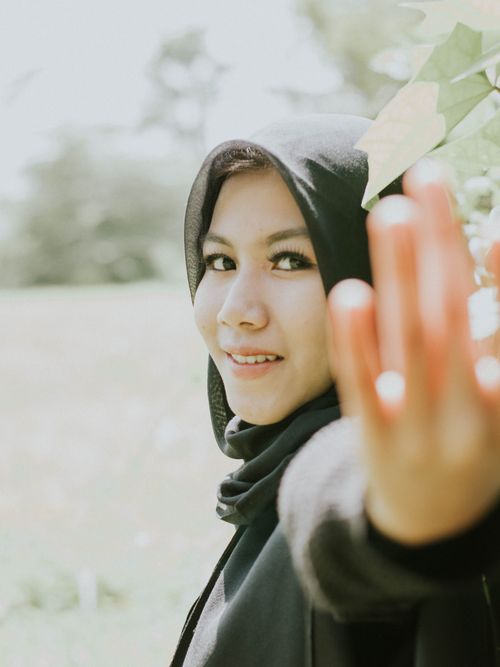 Free A cheerful woman in a hijab smiles brightly while reaching out her hand in a sunny outdoor scene. Stock Photo