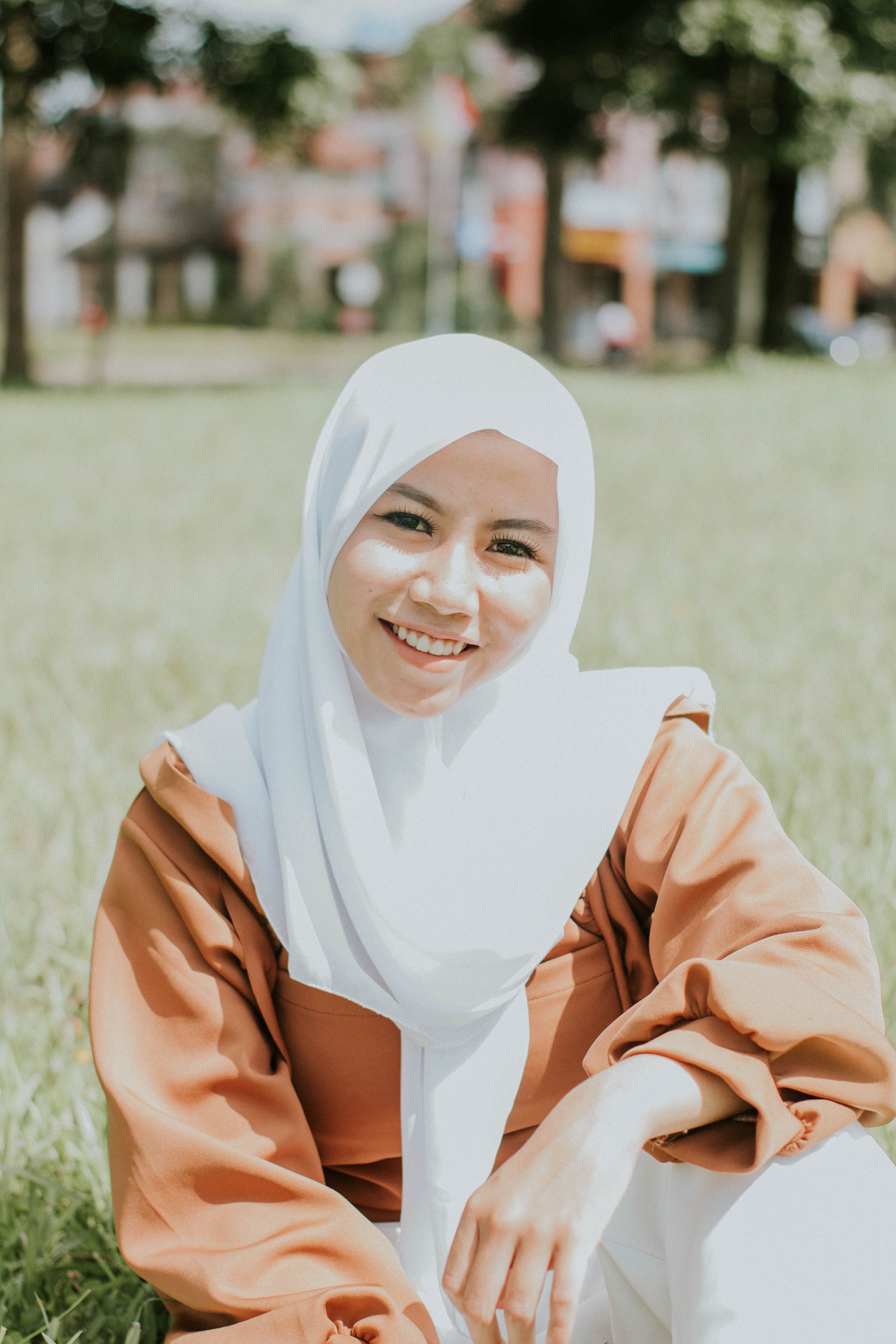 Smiling woman in a hijab sits in a sunlit field, exuding warmth and happiness.