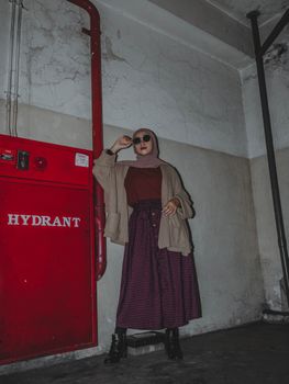 Fashionable woman posing indoors by a hydrant with sunglasses and a hijab, in warm autumn tones.