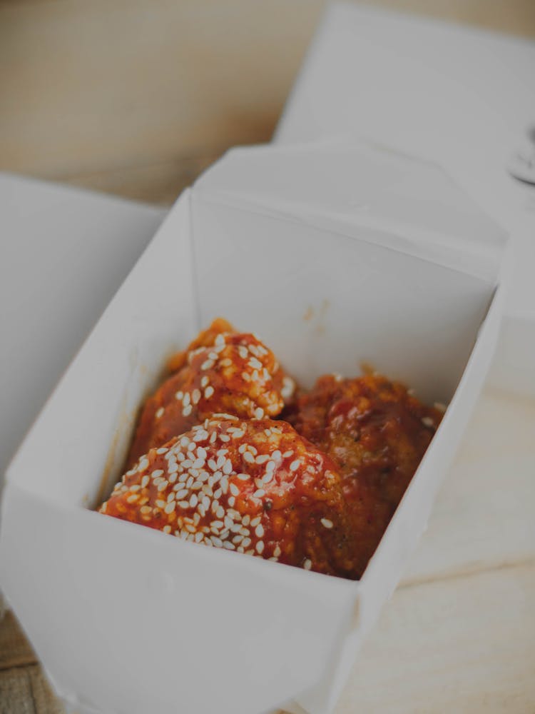 A Take Out Box With  Spicy Chicken Topped With Sesame Seeds