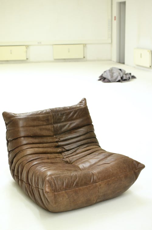 Interior of light spacious room with brown leather pouf on white floor near gray pile of clothes near walls with radiators