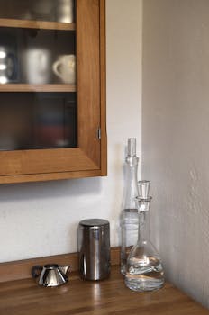 Light kitchen with wooden counter with glass bottles with water and pots near cabinet with mugs on shelves on white wall