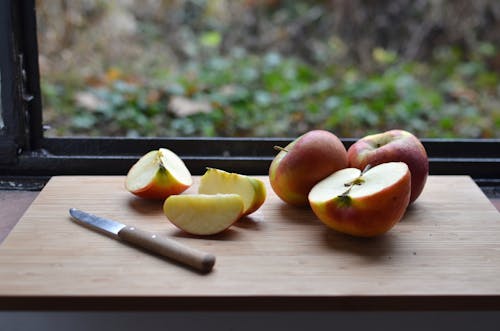 High angle of cut red apples placed on wooden cutting board with knife near window