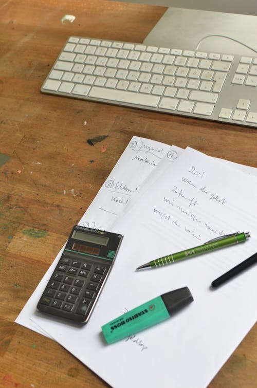 High angle of papers with notes placed with calculator and pens near computer at workplace