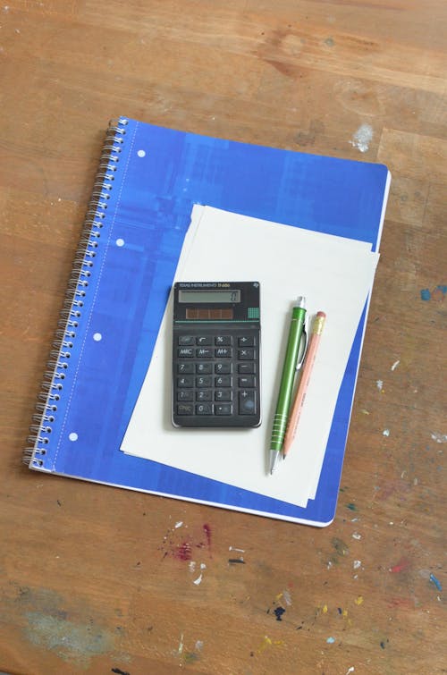 Overhead of pen and pencil near calculator placed on notebook with blue cover and paper sheet on shabby wooden table with paint spots