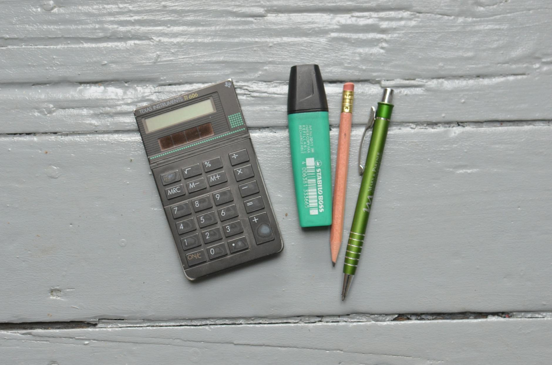 Top view of pen and pencil near green highlighter and calculator placed on old white wooden table
