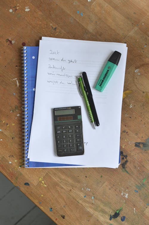 Overhead view of pens and calculator placed on paper sheet with notes on notebook