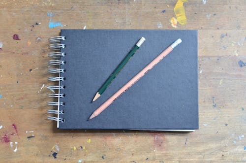 Top view of blank black notepad with pencils placed on shabby wooden surface stained with colourful paints