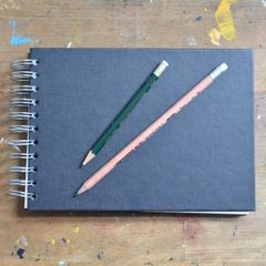A dark spiral-bound sketchbook with two pencils on a paint-splashed table