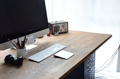 Modern computer with keyboard and portable radio on wooden table near white wall in light comfortable workspace