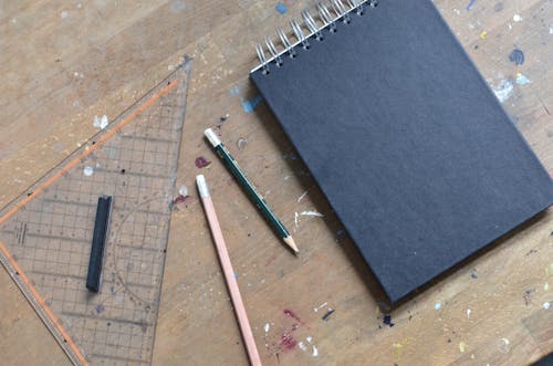 Top view of empty opened notebook with black sheets and pencils near triangular ruler placed on shabby wooden table with paint spots