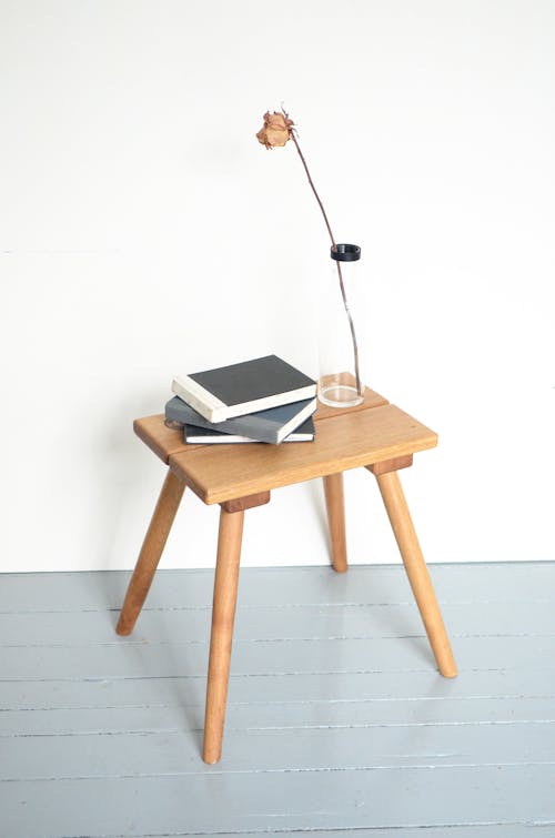 High angle of glass vase with dry rose composed with pile of books on small wooden table in minimalist apartment