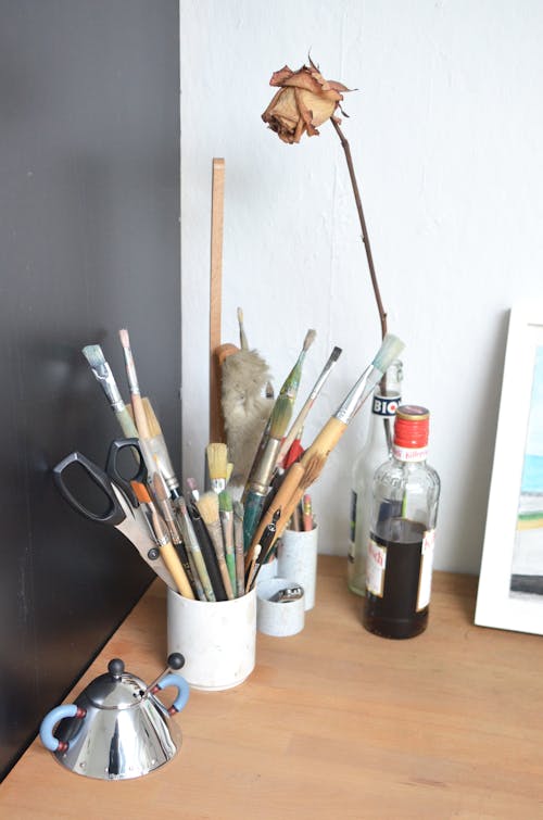 From above of various paintbrushes with scissors and pencils placed in cup arranged on wooden table near dry rose in glass vase in creative art studio