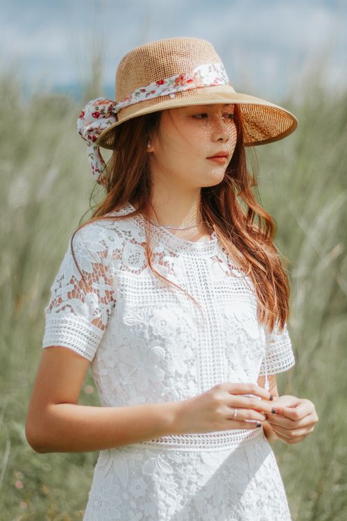 Free Thoughtful Asian female wearing elegant dress and straw hat standing among tall grass in countryside Stock Photo