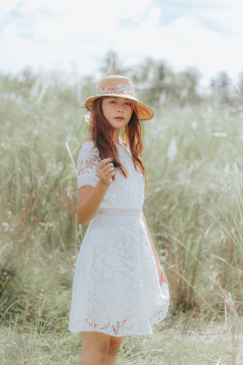 Free Peaceful Asian female wearing straw hat and sundress standing in grassy meadow and looking at camera Stock Photo