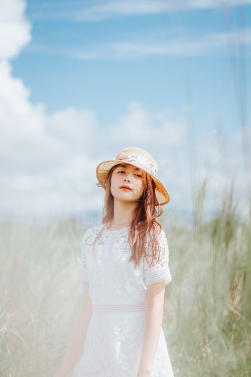 Free Charming Asian female in white dress and straw hat standing in grassy terrain Stock Photo