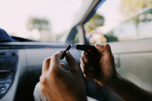 Hands holding a lighter and cannabis joint inside a car, outdoors.