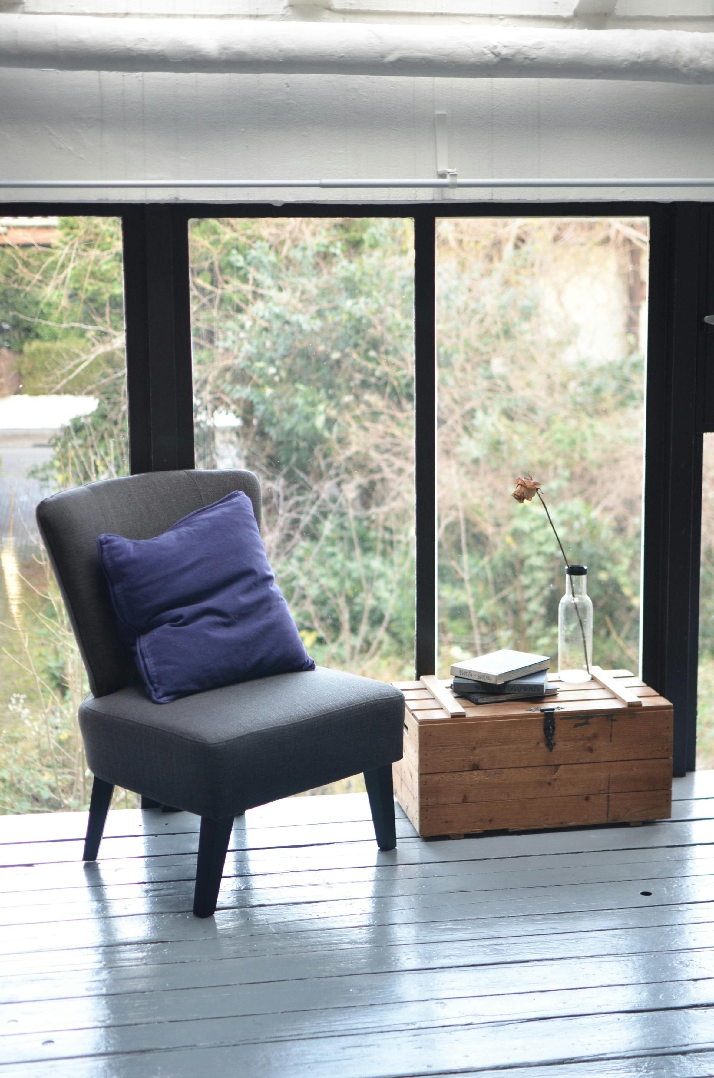 Comfortable gray armchair with blue cushion near books and bottle with flower on wooden box placed in modern light apartment with full wall window