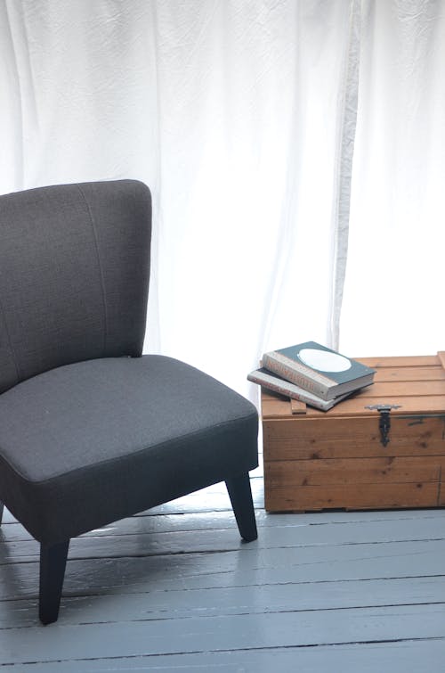 Comfortable gray seat with soft cushion standing on blue wooden floor near nightstand with books on top in light room