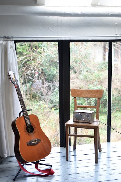 Guitar standing near chair in front of window with radio set in apartment