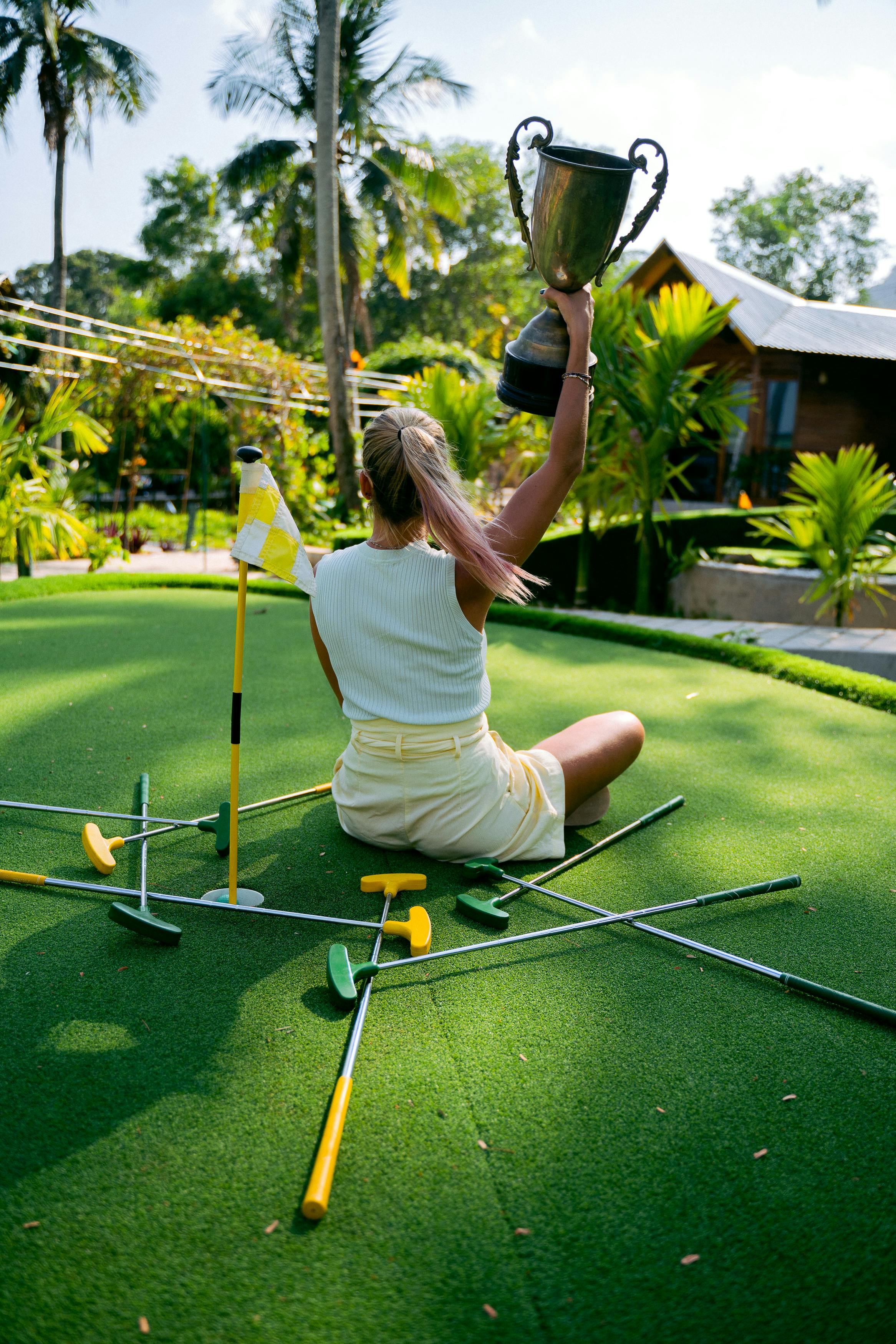 A woman triumphantly raises a trophy while sitting on a lush mini golf course surrounded by putters.