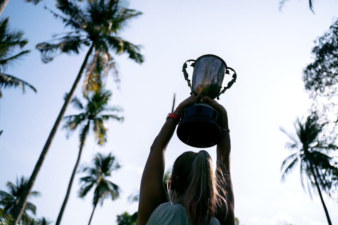 Free A woman lifts a trophy under palm trees, celebrating victory in nature. Stock Photo