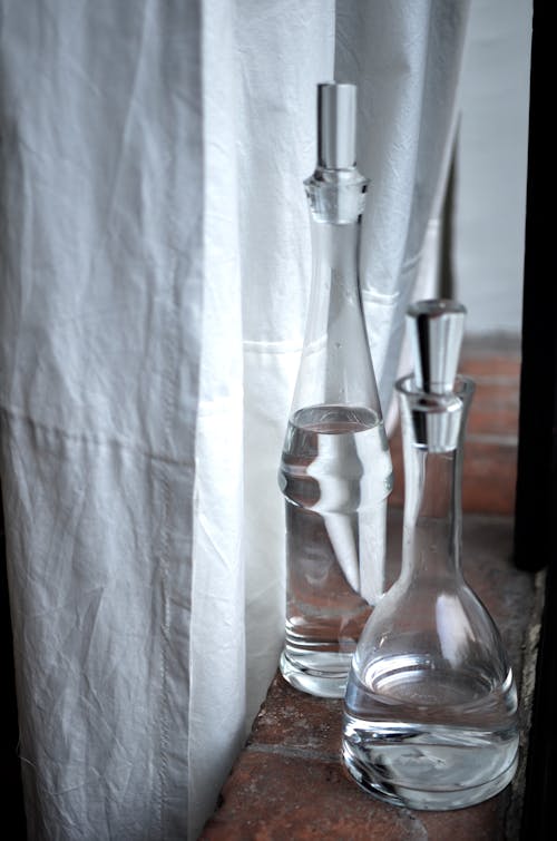 Transparent glass jugs with pure water placed on brick windowsill near white curtain in daytime in room