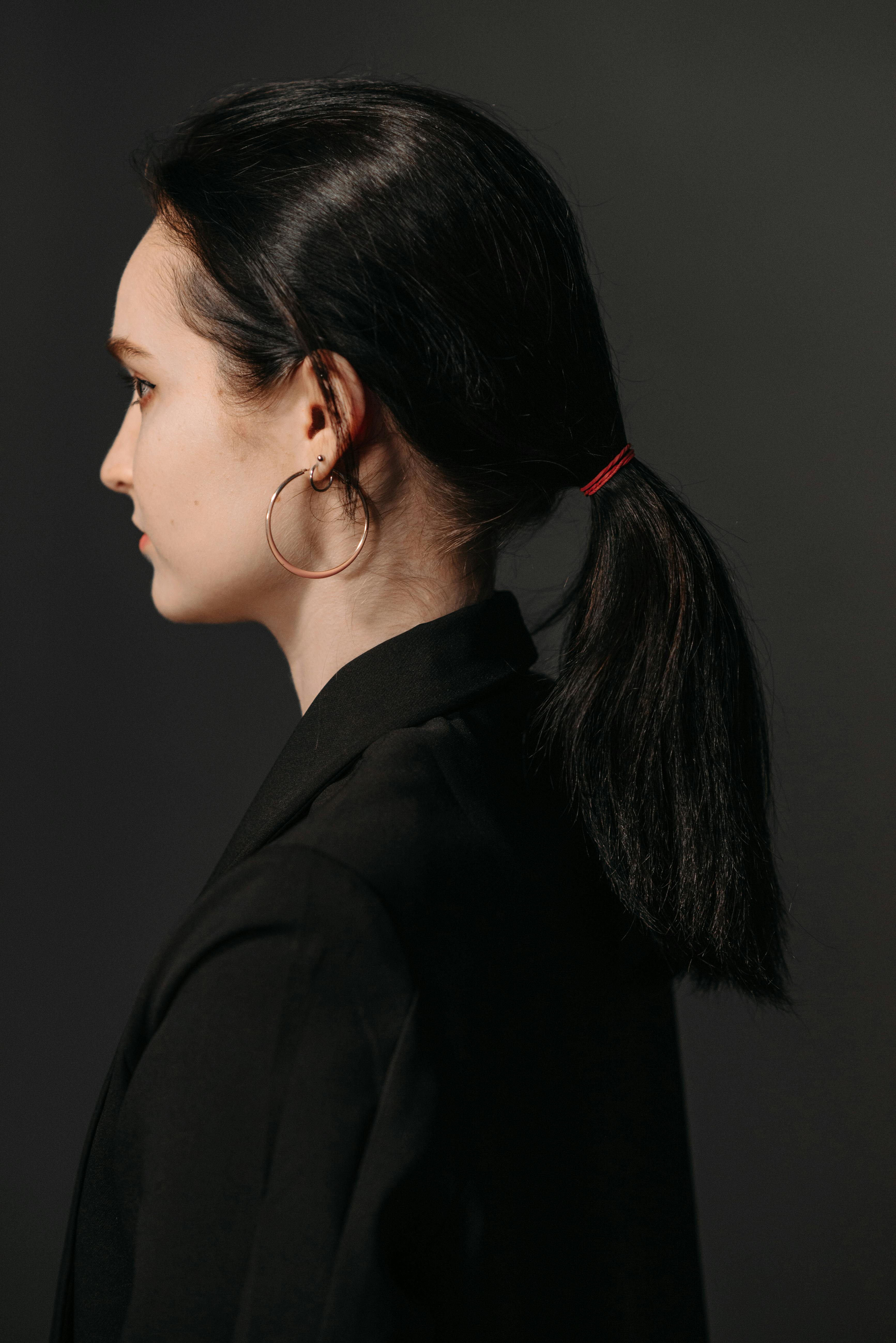 Studio portrait of a woman side profile with tied hair, wearing hoop earrings against a dark background.