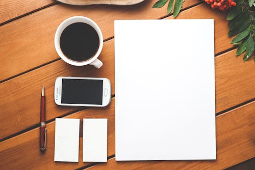 Top view composition of modern smartphone placed near cup of aromatic americano and surrounded with stationeries including blank A4 format paper pen and business card size papers on wooden table