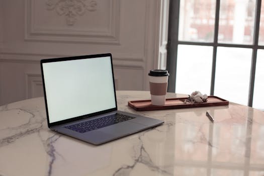 A minimalist interior setup featuring a laptop, coffee cup, and pen on a marble table by a window.