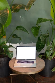 Modern laptop with blank screen on table, framed by large green plants, ideal for mockups.