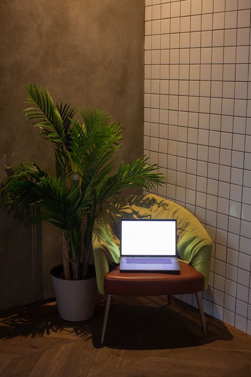 Free A cozy indoor workspace featuring a sleek laptop on a chair beside a potted plant against tiled walls. Stock Photo