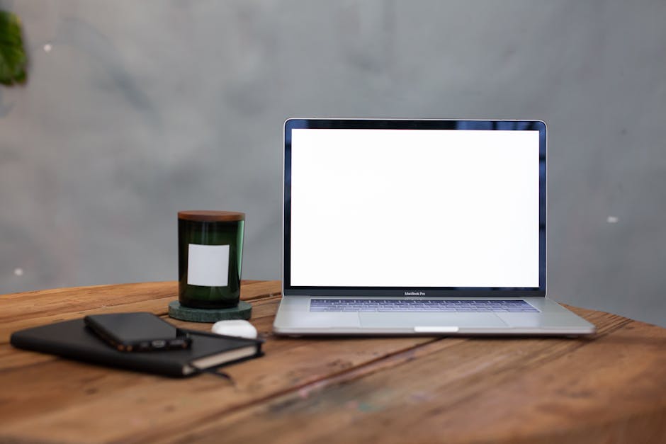 A modern workspace featuring a blank laptop screen, notebook, and candle on a wooden table.