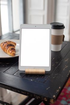Tablet on a stand with empty screen next to a croissant and coffee on a vintage table.