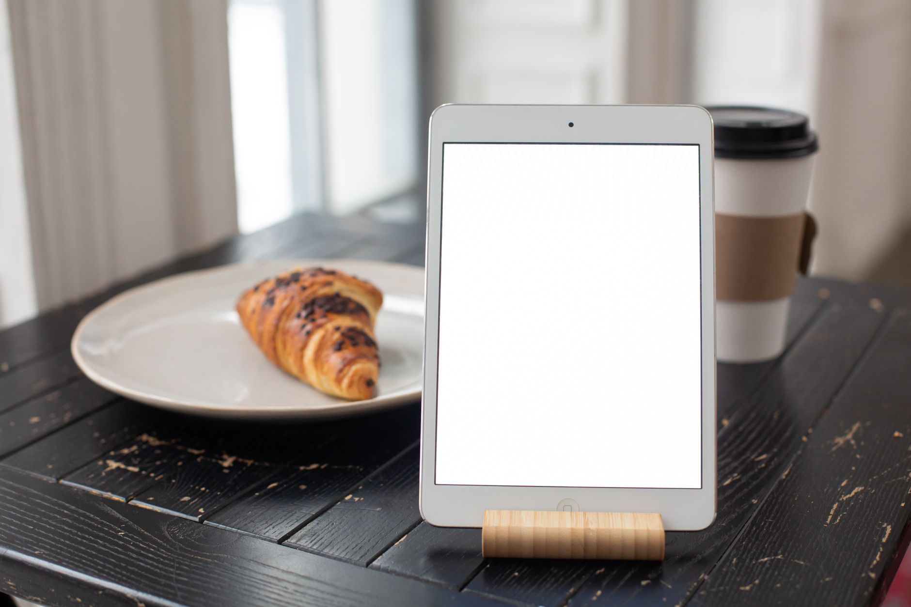 A blank tablet on a wooden stand with a coffee and croissant on a rustic table.