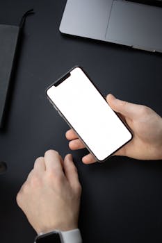 Hands holding a smartphone with blank screen over a black desk, perfect for mobile-related themes.