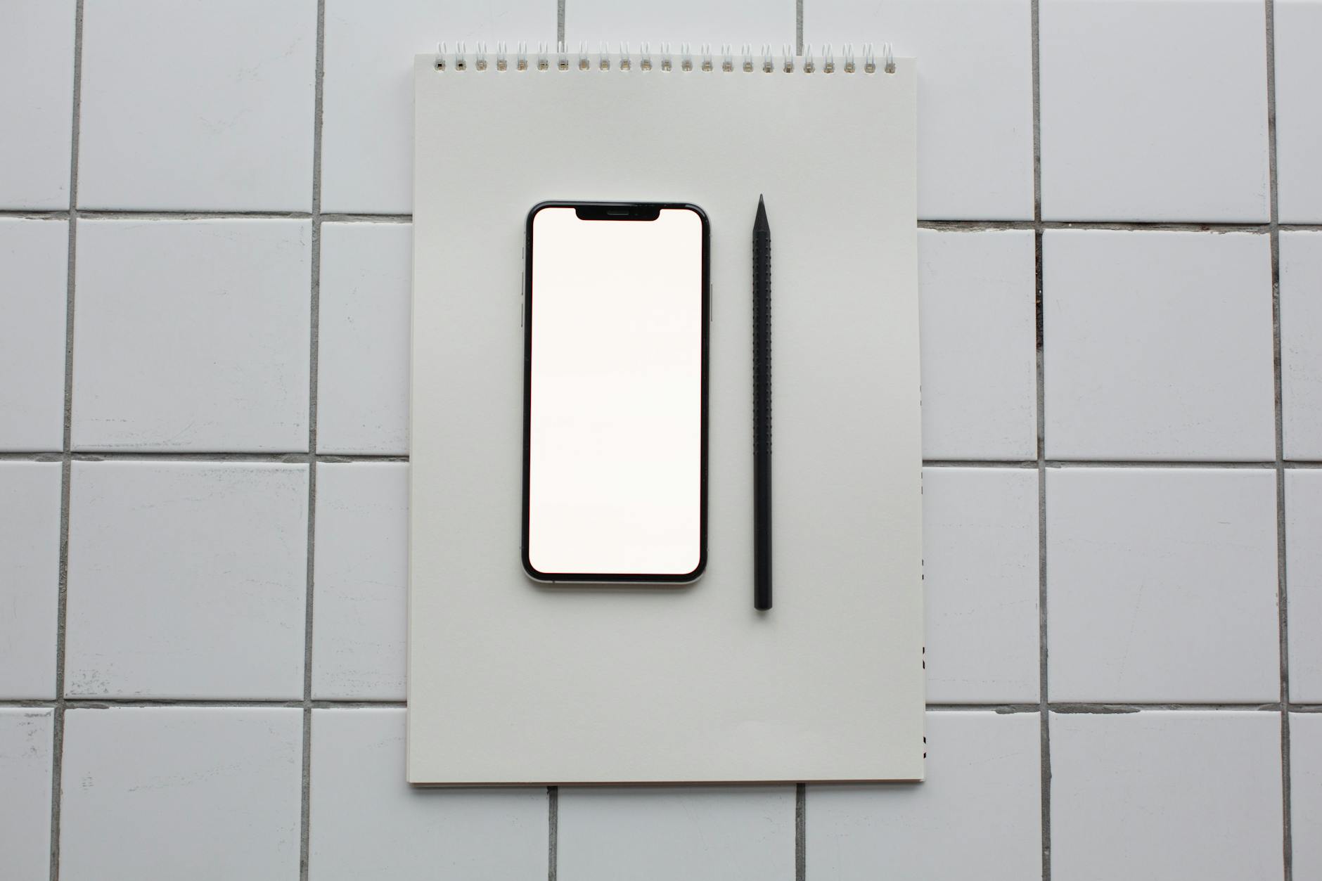 Flat lay of a smartphone with blank screen on a notebook next to a pencil, on a tiled surface.