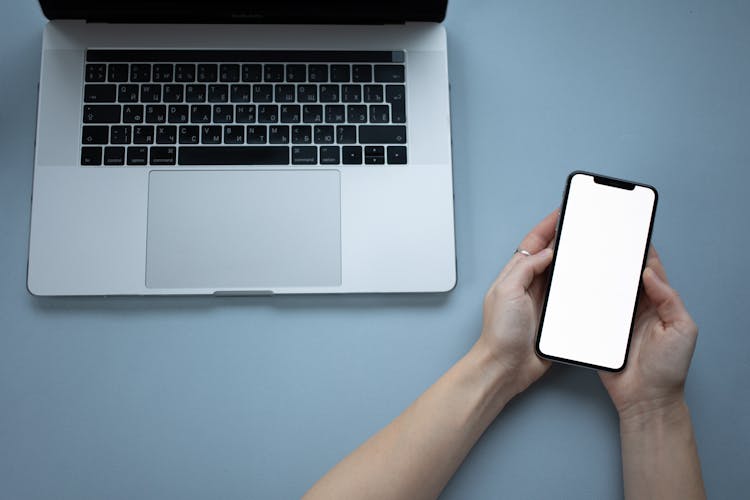 A Person Holding A Smartphone With Blank Screen Near A Laptop
