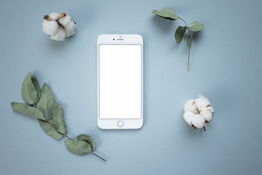 Flat lay of a smartphone surrounded by cotton and leaves on a blue surface, ideal for tech and nature themes.