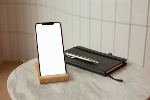 A smartphone with blank screen on a wooden stand beside a notebook and pen, indoors.