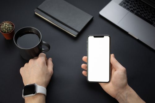 Free A minimalistic workspace setup featuring a smartphone, coffee mug, and smartwatch on a dark desk. Stock Photo