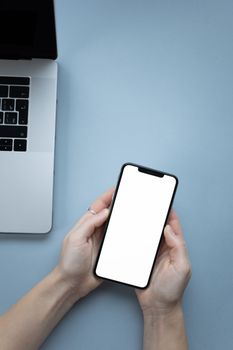 A person holding a blank smartphone above a laptop on a blue surface, ideal for mockup use.