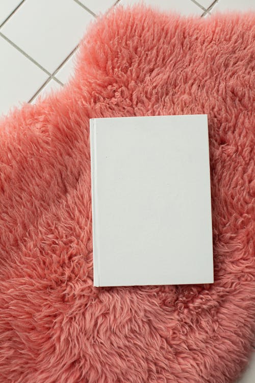 From above of notebook with white blank cover placed on pink shaggy rug on tiled floor