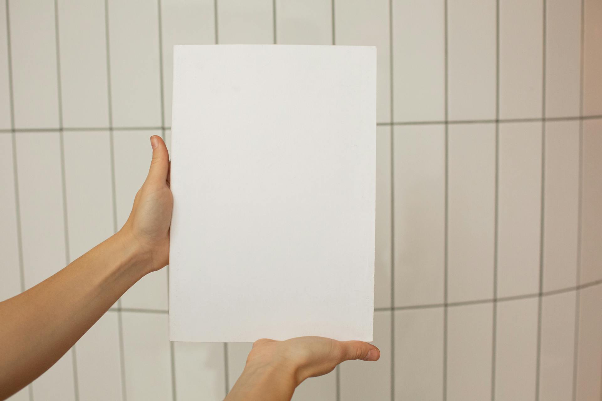 Crop faceless person showing empty sheet of paper with tiled wall on background