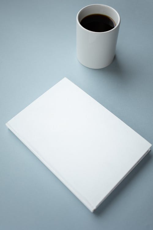 High angle of mockup of white hardcover of notebook placed near white ceramic cup of americano on table